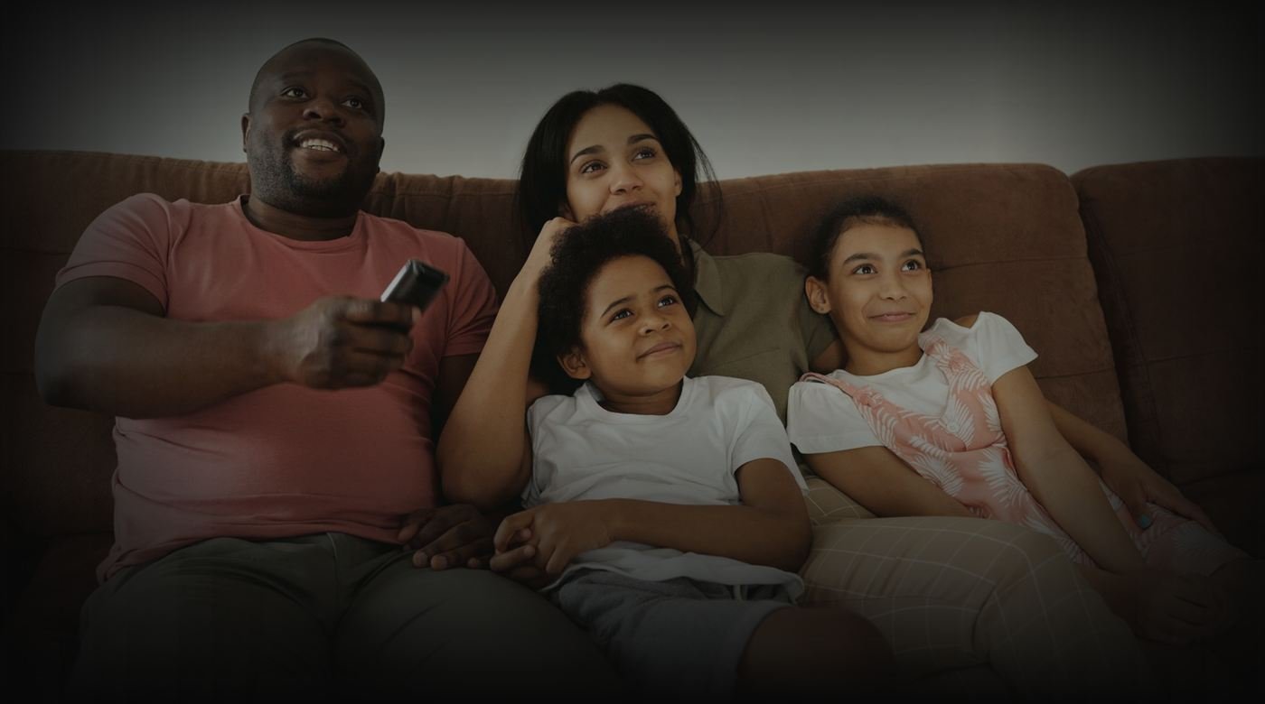 Uma família sentada no sofá da sala assistindo à televisão. O pai está com o controle remoto na mão e as crianças estão encostadas na mãe.